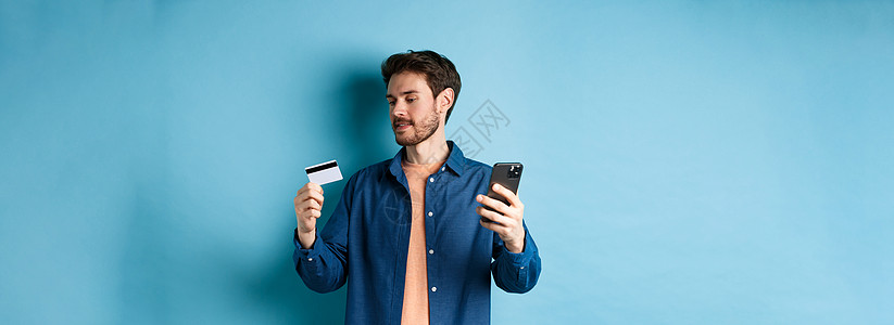 电子商务的概念 男子支付在线支付 读取信用卡号码并手持手机 站在蓝色背景上情绪工作室发型技术成人电话男人成功胡子胡须图片