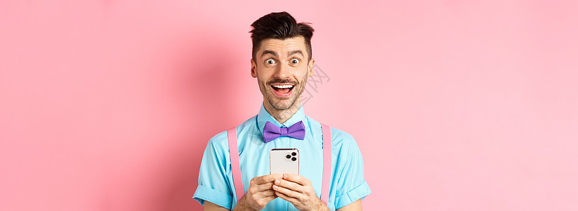 阅读智能手机屏幕 在相机上微笑 站在粉红背景面前的欢笑和兴奋的人 他们眼中的喜悦之人工作室广告发型胡子胡须快乐成人办公室吊带裤男图片