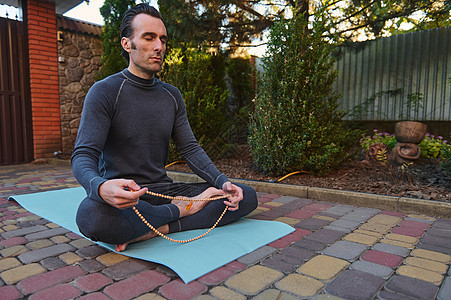 和平的瑜伽男子 坐在健身垫上摆着莲花 在户外做瑜伽练习时与玫瑰珠子一起冥想图片