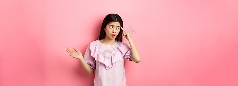 你傻吗 震惊的亚洲女性指着头抱怨 骂某人疯了或奇怪 看向一边 站在粉红色背景下青少年学生工作室促销情感伙伴投标化妆品购物女士背景图片