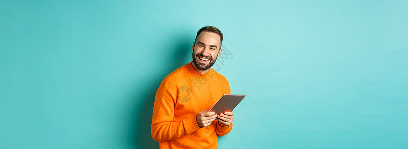 英俊的长胡子男子使用数字平板电脑 嘲笑照相机 在浅蓝背景下快乐地站立学生工作室橙子购物促销男性横幅药片黑发男人图片