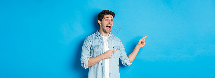 英俊男人的肖像 看着并用手指指着左手 兴奋地站在蓝色背景上快乐男性潮人成功手势广告闲暇胡子技术标识图片