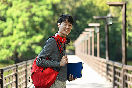 照片中笑着的亚裔男子大学生 手拿着背包和书本站在河上桥上图片