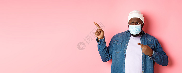 Corona病毒 生活方式和全球大流行病概念 脸部蒙面的迷糊和失望的非裔美国人用左手指着手指 盯着摄影机 粉红色背景隔离男人快乐图片