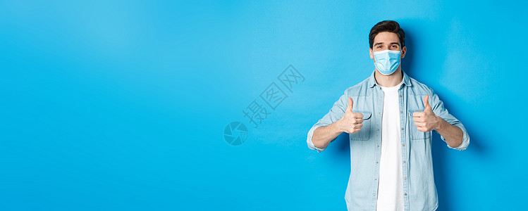 身戴医疗面具的年轻人在蓝背景下站立 表现出赞同 同意和认同的拇指举起脚趾头ACN 9WG IWP促销黑发隔离情绪成功广告口罩商业图片