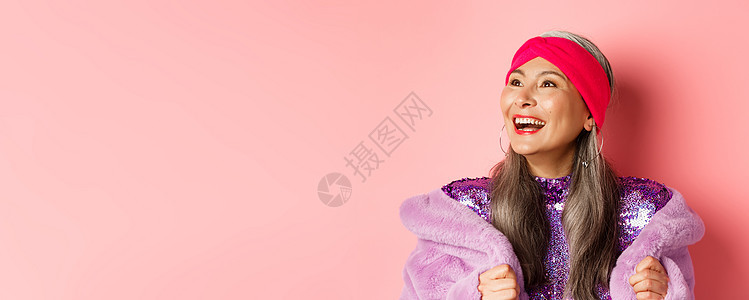 时装 穿着紫色的毛皮大衣 粉红背景 站在左面看着快乐的脸脸朝左边笑着祖母奶奶广告老年头发头巾购物裙子女性乐趣图片