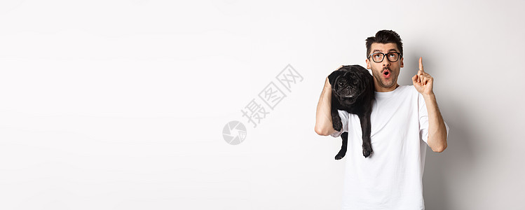 戴着眼镜的帅帅帅壮男子 肩上抓着可爱的黑毛狗 用宣传标志指尖 站在白色背景之上微笑横幅朋友黑色潮人工作室行动宠物犬类男性图片