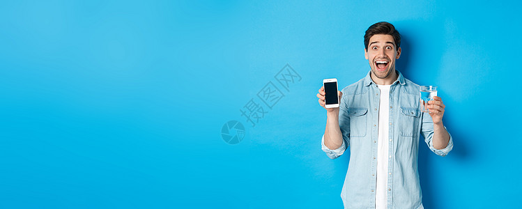 快乐的男子展示水杯和移动屏幕 建议智能手机健康应用程序 站在蓝色背景面上   info whatsthis情绪黑发商业互联网技术图片