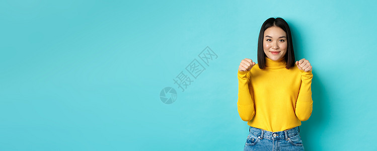 美丽和时尚的概念 穿着黄色套头衫的美丽时尚的亚洲女性 双手举在胸前 仿佛拿着横幅或标志 站在蓝色背景上发型成人商业广告女孩工作室图片