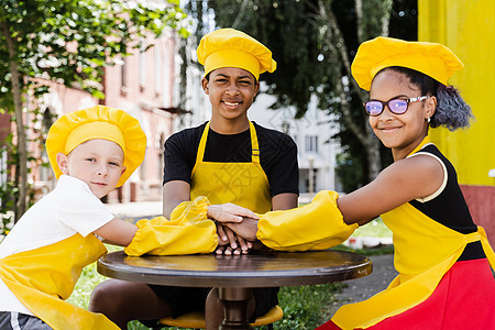 多种族儿童一起做饭 手牵着手堆成一堆 跨国孩子的友谊 童年 身着厨师帽 身着黄色围裙制服的厨师们把手放在彼此身上姐姐食物面粉面团图片