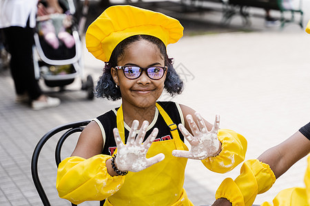 黑非洲厨师女孩用面粉和微笑举手示众 非洲儿童穿着厨师帽和黄色围裙制服图片