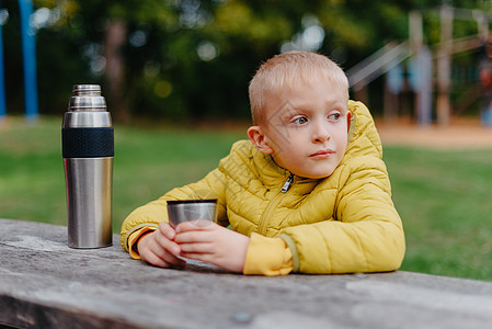 男孩坐在木桌旁 端着热水瓶和一杯茶 望着远方 秋天 一个男孩坐在公园的一张桌子旁 儿童男孩儿子在秋天公园 坐木凳和桌子 户外的小图片