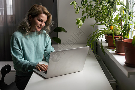 穿着休闲蓝色衬衫的年轻微笑快乐满意的员工女商务人士拿着笔坐在工作场所的白色办公桌前工作 在室内轻便的现代办公室里拿着笔记本电脑 图片