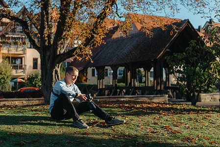 专业摄影师拍摄美丽的秋天公园 男子专业摄影师坐在秋天公园的相机和智能手机 润饰 鲜艳的色彩 褐色调女士男人森林男性艺术家工作电话图片