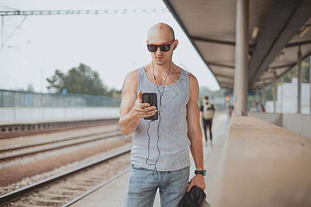 穿着 T 恤的年轻人在站台上用手机等火车 男子在郊区火车站站台旁 提着包和手机 乘火车旅行运输平台游客车站铁路幸福通勤者背包民众图片