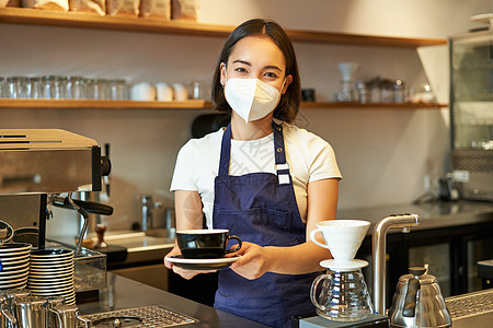 Covid 19 和热情好客 戴着医用面具微笑的亚洲女咖啡师 给客户一杯咖啡 在咖啡馆柜台后面准备订单 穿着制服服务学生顾客商务图片