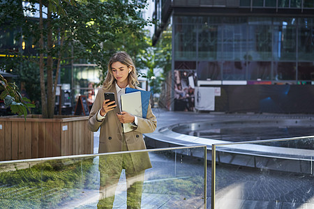 穿着西装的企业女性肖像使用手机 在市中心等待商业伙伴 拿着工作文件和笔记本电脑电话员工公司女士代理人街道人士城市售货员套装图片