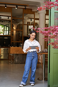 年轻的有平板药的亚洲女性 站在咖啡厅入口前 邀请客人和顾客 对着镜头大笑调酒师人士商务咖啡经理咖啡师商业自雇酒吧女孩图片