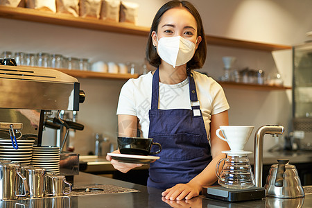Covid 19 和热情好客 戴着医用面具微笑的亚洲女咖啡师 给客户一杯咖啡 在咖啡馆柜台后面准备订单 穿着制服服务员女性女士咖图片