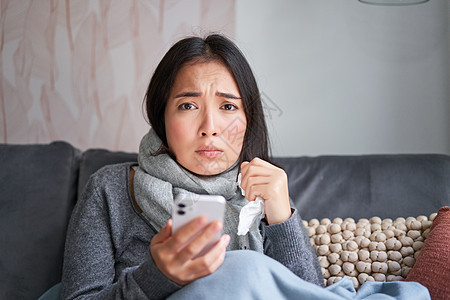 患有流感的亚洲女孩 坐在家里 哭泣 看起来很沮丧 拿着智能手机 感觉不舒服 阴沉 穿着暖和的衣服鼻子温度长椅弱点温度计过敏药品沙图片