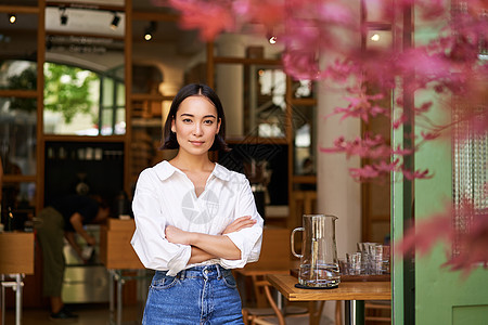 站在楼顶年轻女商务人士在她自己的咖啡馆的肖像 经理站在入口近处邀请你 穿着白光衬衫和牛仔裤酒吧食堂女孩咖啡师自雇调酒师员工人士横幅互联网背景