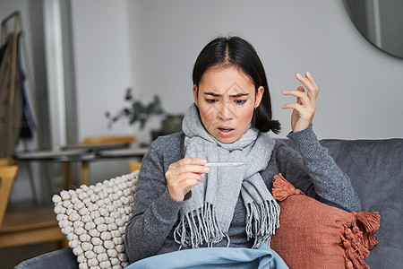 惊吓的女人看着她的温度计 感寒 发烧 坐在家中休病假等毯子沙发女士女性鼻子疼痛病人流感症状互联网图片