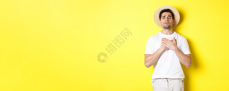 旅游和夏季的概念 戴着草帽的浪漫男人看起来怀旧 闭上眼睛 手放在心上 站在黄色背景下假期手势微笑闲暇商业游客情绪潮人工作室旅行图片