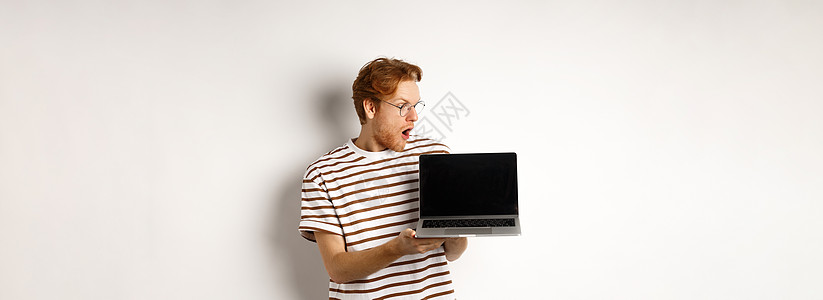 红发年轻人看起来印象深刻 展示了笔记本电脑空空屏幕上标志横幅的标语 Guy在眼镜中检查计算机 白背景在线广告工作自由职业者男性胡图片