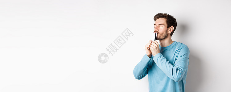 年轻男子亲吻塑料信用卡时面满脸满意 穿着蓝轻便衬衫站在白色背景上站立商业胡子银行工作室生活发型胡须成功情绪促销图片