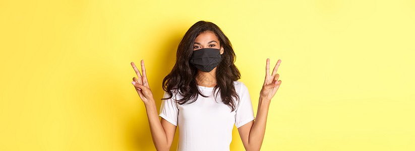 covid19 的概念 社会距离和生活方式 戴着黑面罩的美丽非洲裔美国女孩的画像 站在黄色背景上 展示着和平标志 微笑着疾病情感图片