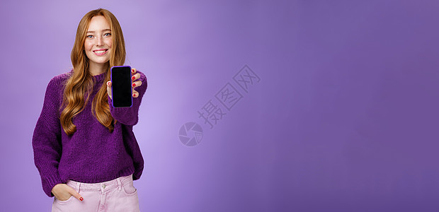 女孩在相机前展示智能手机屏幕 询问朋友的意见 面带乐观和快乐的表情 手牵着口袋 在紫色背景下推销手机或应用程序电话毛衣雀斑寒冷互图片