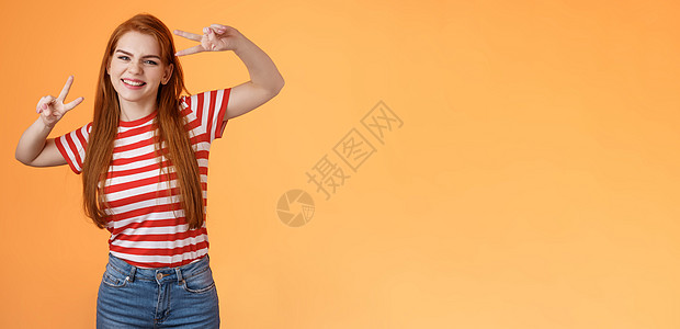 友好乐观的漂亮红发女人欢欣鼓舞 感到快乐 享受夏日时光 拍第一张照片假期 给同事发夏日照片 制作和平胜利标志 微笑着宽阔的橙色背图片