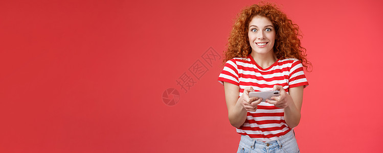 兴奋快乐可爱的红发卷发女友急切地开始学习如何玩男朋友游戏看起来很有趣快乐的相机微笑快乐持有水平智能手机加载应用程序红色背景理发社图片