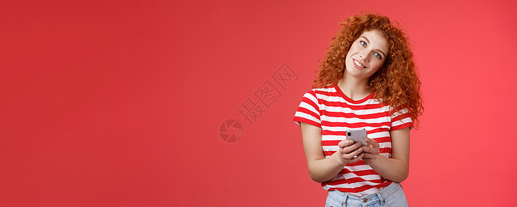 愚蠢可爱的红发卷发时尚女性拿着智能手机倾斜头微笑着露出露齿的正面笑容思考什么答案发送挑衅的时髦信息红色背景青年电话展示媒体技术社图片