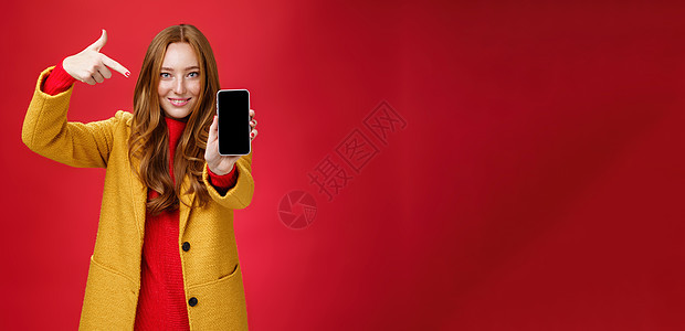选择这款您永远不会后悔的手机 身穿黄色外套 长相友善 自信的红发魅力女友的肖像 智能手机指着手机 对着镜头微笑互联网女性雀斑工具图片