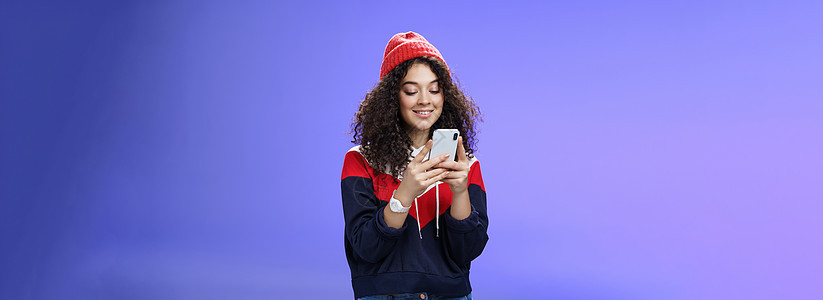 在互联网搞笑模因中搜索的女性 手持手机 在智能手机屏幕上开心微笑 可爱 在蓝色背景下打字或浏览互联网 戴着温暖的冬帽女孩手表工具图片