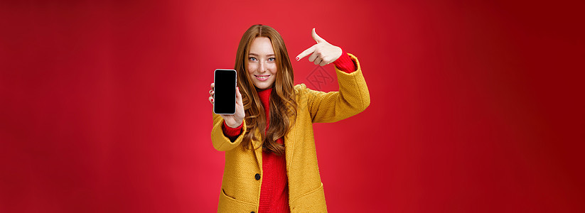 选择这款您永远不会后悔的手机 身穿黄色外套 长相友善 自信的红发魅力女友的肖像 智能手机指着手机 对着镜头微笑喜悦成人新年发型工图片