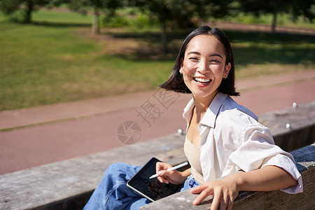 带着数字平板和图形笔坐在长椅上 笑着微笑 对着镜头转过身来并热烈说话的韩国快乐妇女艺术家女孩公园享受福利药片黑发绘图喜悦治疗图片