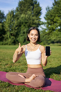 对亚洲女性的垂直拍摄显示拇指向上 建议在线瑜伽训练 冥想应用 在公园新鲜空气中做操练 智能手机屏幕女孩日落运动成人女士赛跑者慢跑图片