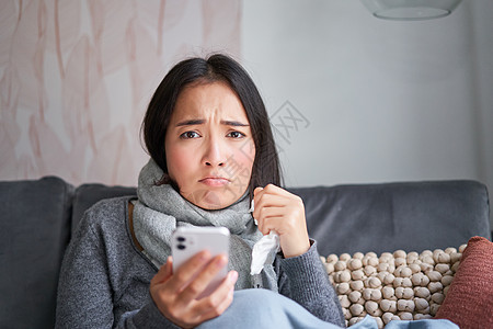 患有流感的亚洲女孩 坐在家里 哭泣 看起来很沮丧 拿着智能手机 感觉不舒服 阴沉 穿着暖和的衣服女性组织鼻子互联网药品温度计温度图片