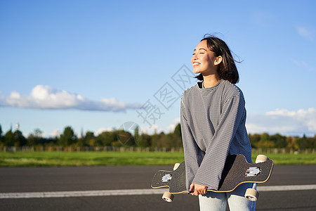 韩国女孩滑冰 手握滑板 在路上摆姿势 微笑和看一眼 笑着笑着溜冰者木板运动女性乐趣闲暇行动太阳潮人青少年图片