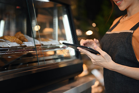 微笑的咖啡店老板使用数字平板电脑接收在线订单 同时站在展示蛋糕和甜点的橱窗旁图片