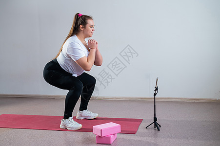 一个胖胖的年轻女子正在看一个在线健身课 在手机上 远程体育培训健身房运动装手表练习会议班级腹肌健康讲师运动图片