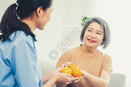 一位年幼的照顾者将橘子递给家里她满足的老年病人老年学护理看门人药品工人长老服务医学女性医生图片