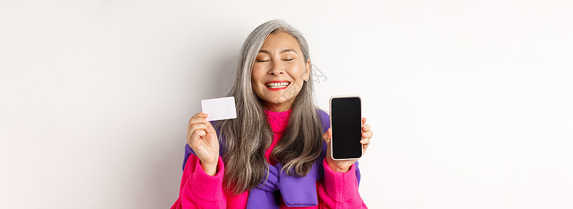 欢乐和快乐的亚洲年长妇女微笑 展示空白智能手机屏幕 塑料卡 互联网购物 白背景等信息  Twitter毛衣时尚促销奶奶派对广告化背景图片