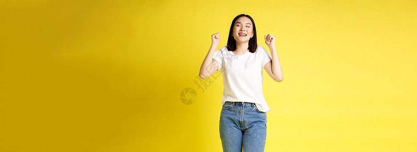 穿着牛仔裤和白色 T 恤的快乐亚洲女性的全尺寸照片从幸福中跳出来 赢得奖品和欢欣鼓舞 欢呼雀跃 欢呼雀跃 黄色背景黑发成就青少年图片
