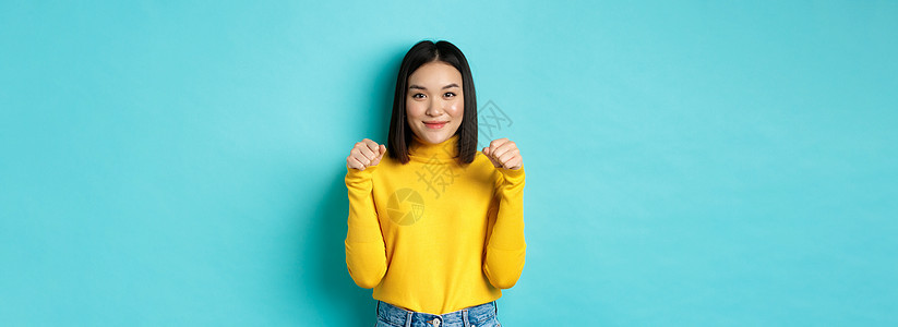 美丽和时尚的概念 穿着黄色套头衫的美丽时尚的亚洲女性 双手举在胸前 仿佛拿着横幅或标志 站在蓝色背景上发型情感黑发学生成人促销青图片