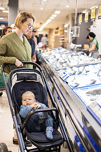 穿着随意的母亲在超市杂货店的鱼市部选择鱼 带着她的婴儿男孩在婴儿车里孩子父母妈妈市场销售零售产品海鲜生物食物图片