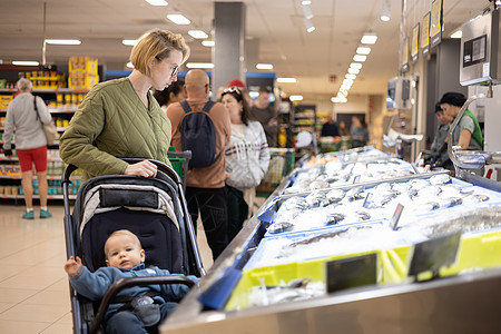 穿着随意的母亲在超市杂货店的鱼市部选择鱼 带着她的婴儿男孩在婴儿车里儿子服务产品店铺妈妈购物中心顾客零售海鲜孩子图片