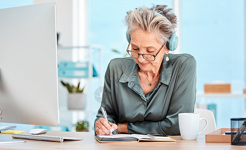 高级女性 在办公桌上戴着音乐耳机写作和安排时间 阅读或专注 年长的秘书 接待员或通信主管在桌上用笔记本 个人电脑或听收音机图片